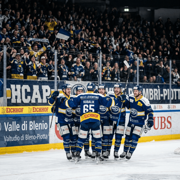 Six HC Ambrì-Piotta ice hockey players in blue uniforms celebrating on ice with cheering crowd behind